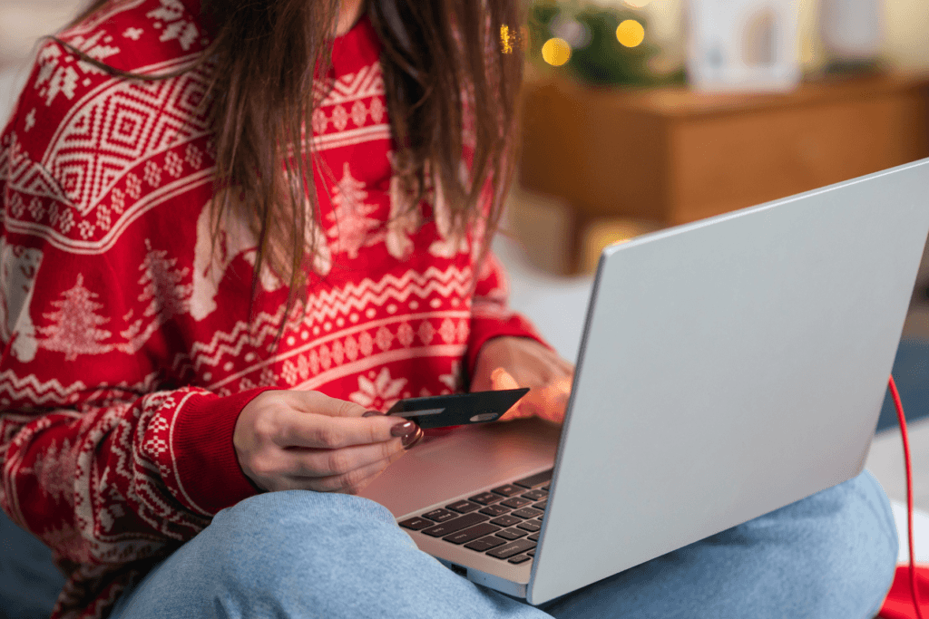 A girl in a red holiday sweater holds a credit card while using a laptop to depict holiday online shopping